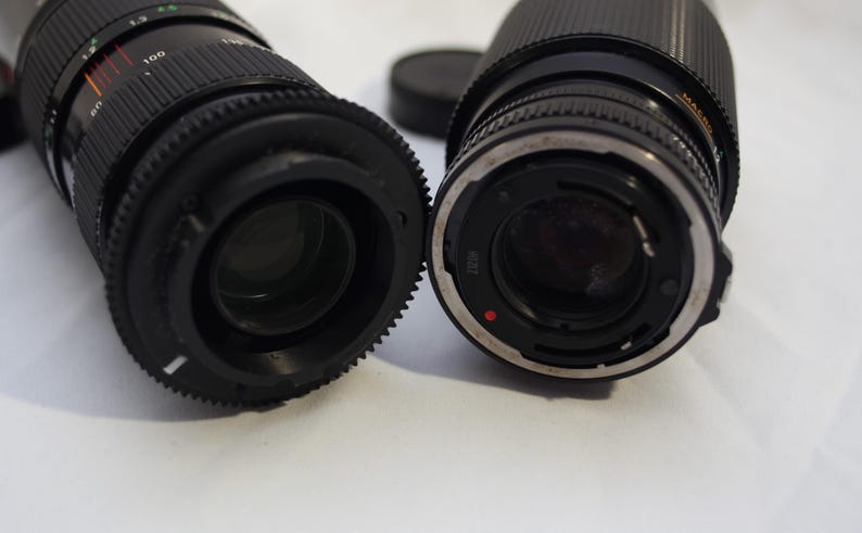May include: Two black camera lenses with silver accents. The lens on the left has a black and red ring with the number 100 printed on it. The lens on the right has a silver ring with a red dot.