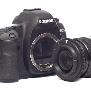 May include: Black Canon EOS 5D camera body with a detached lens. The camera has a textured grip and an orange strap. The lens is black with measurement markings. The Canon logo is visible on the camera body.