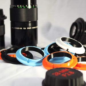 May include: A collection of camera lens adapters in various colours, including black, white, blue, and orange. The adapters are made of plastic and have a smooth, glossy finish.