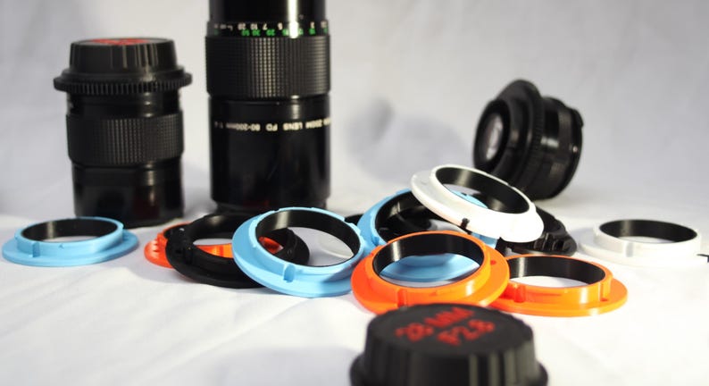 May include: A collection of camera lens adapters in various colours, including black, white, blue, and orange. The adapters are made of plastic and have a smooth, glossy finish.