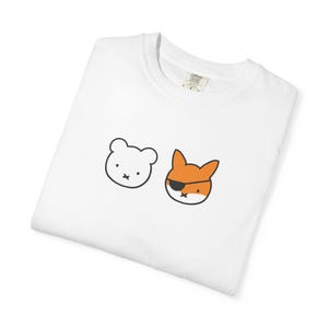 Könnte beinhalten: Ein weißes T-Shirt mit Rundhalsausschnitt und einer minimalistischen Grafik von zwei Tierköpfen. Links ist ein weißer Bärenkopf mit schwarzer Umrandung zu sehen, rechts ein orangefarbener Fuchskopf mit schwarzer Augenklappe und schwarzer Umrandung.
