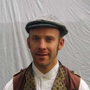 May include: A gray tweed flat cap. The cap is worn with a white collared shirt and a brown vest with a patterned design. The vest has brown shoulder accents and is buttoned up. The background is a light gray.