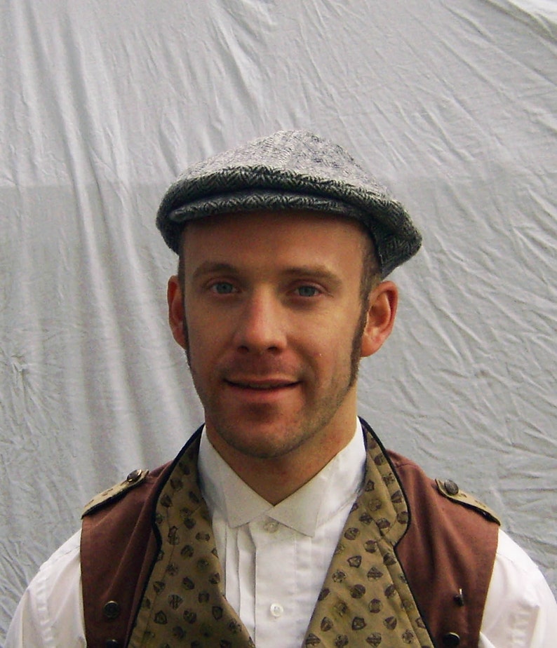 May include: A gray tweed flat cap. The cap is worn with a white collared shirt and a brown vest with a patterned design. The vest has brown shoulder accents and is buttoned up. The background is a light gray.