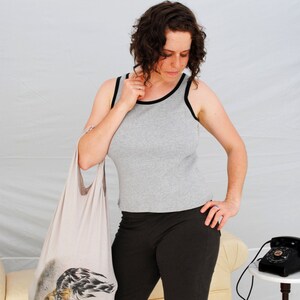 以下が含まれることがあります： グレーのタンクトップに黒いトリムと黒いショートパンツを着用した女性が、白地に黒と白のグラフィックプリントが施された白いトートバッグを持っています。彼女は白い背景の前に立っています。