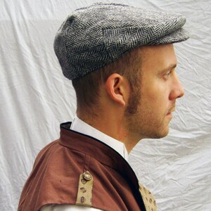 May include: A gray and white herringbone pattern newsboy cap. The cap has a rounded crown and a small, stiff brim. The cap is made of a textured fabric. The person is wearing a brown and white outfit.