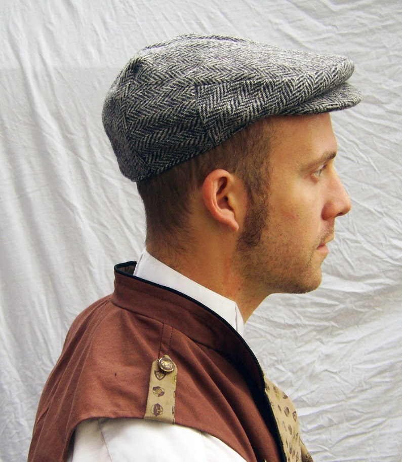 May include: A gray and white herringbone pattern newsboy cap. The cap has a rounded crown and a small, stiff brim. The cap is made of a textured fabric. The person is wearing a brown and white outfit.