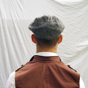 May include: A gray herringbone tweed newsboy cap. The cap is worn by a person wearing a white collared shirt and a brown vest. The vest has gold-colored shoulder accents.