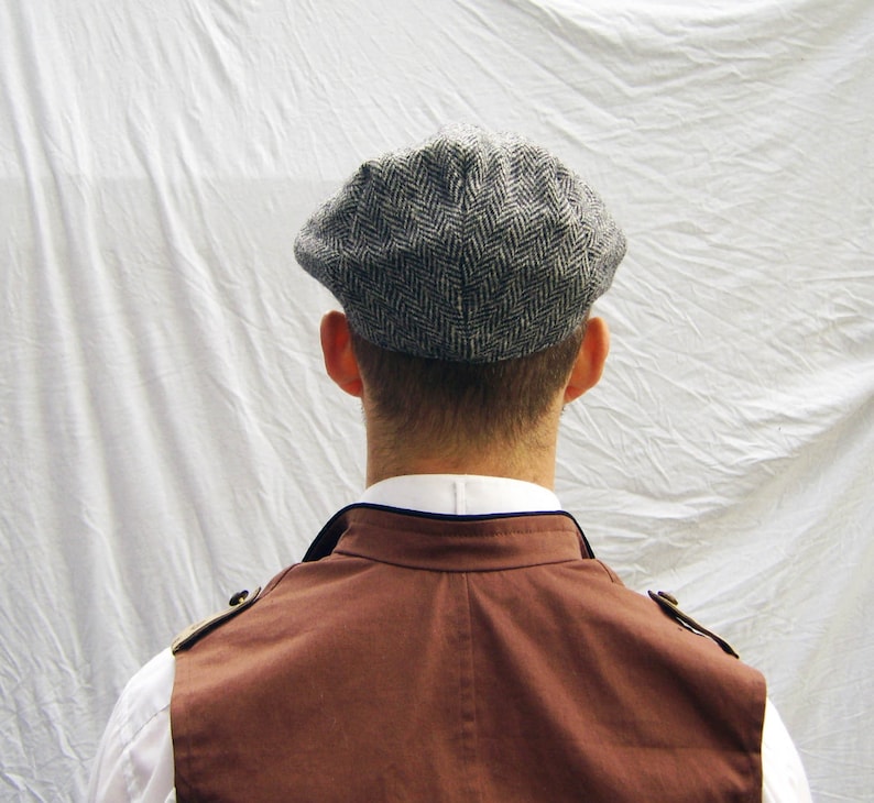 May include: A gray herringbone tweed newsboy cap. The cap is worn by a person wearing a white collared shirt and a brown vest. The vest has gold-colored shoulder accents.