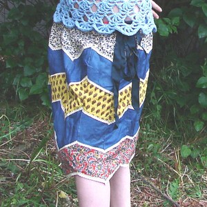 Op de afbeelding: Een blauwe en gele patchwork rok met een zwarte strik om te binden. De rok is gemaakt van verschillende stoffen en heeft een golvend patroon.