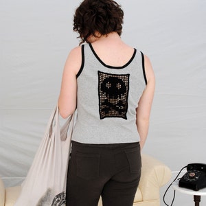 以下が含まれることがあります： 女性は、背中に黒いクロシェの骸骨のデザインが施されたグレーのタンクトップを着ています。彼女は濃い茶色のショートパンツを履いていて、白いトートバッグを持っています。