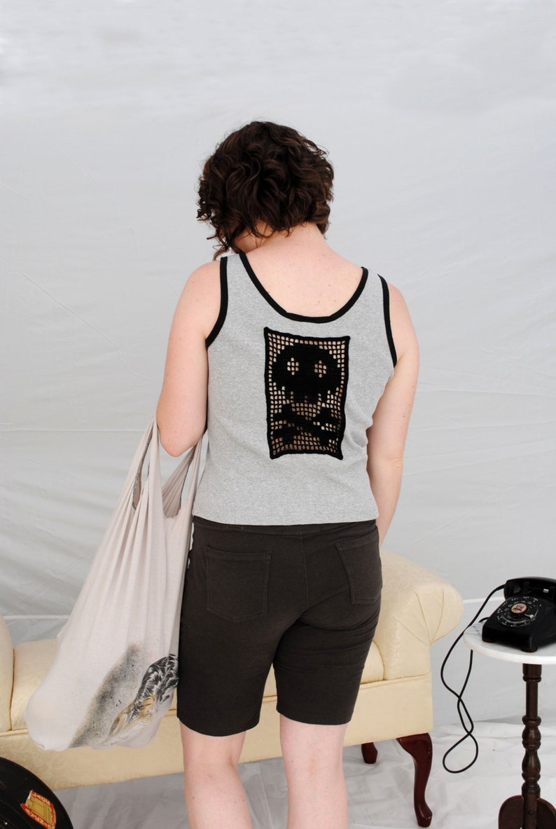 以下が含まれることがあります： 女性は、背中に黒いクロシェの骸骨のデザインが施されたグレーのタンクトップを着ています。彼女は濃い茶色のショートパンツを履いていて、白いトートバッグを持っています。