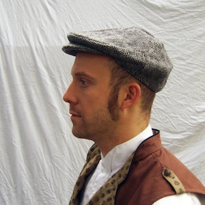 May include: A gray herringbone tweed flat cap. The cap has a rounded crown and a small, stiff brim. The cap is worn with a white collared shirt, a brown vest, and a patterned scarf. The background is a white sheet.
