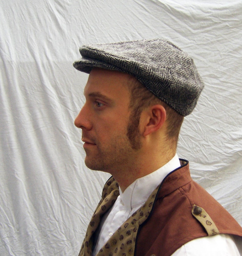 May include: A gray herringbone tweed flat cap. The cap has a rounded crown and a small, stiff brim. The cap is worn with a white collared shirt, a brown vest, and a patterned scarf. The background is a white sheet.