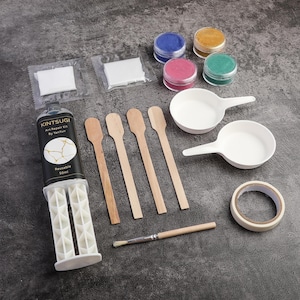 以下が含まれることがあります： 「Kintsugi Art Repair Kit By YanYun Reusable 50ml」と書かれた白黒のラベルが付いた金継ぎアート修復キット。 キットには、木製のかき混ぜ棒、筆、着色粉末、小さな白いボウルが含まれています。