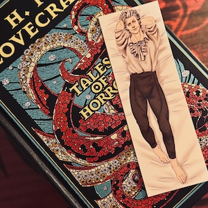 Könnte beinhalten: Ein Lesezeichen mit einem Mann in einem weißen Hemd und braunen Hosen, der auf einem Bett liegt. Das Lesezeichen liegt auf einem Buch mit dem Titel "H.P. Lovecraft Tales of Horror".