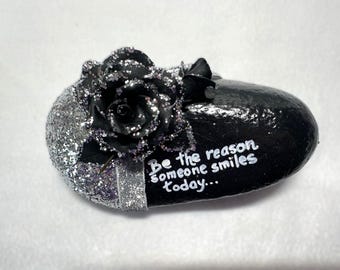 Sé la razón por la que alguien sonríe. Afirmación. Piedra pintada en negro y plata con cinta y flor.