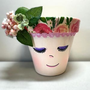 May include: A white ceramic flower pot with a painted face, closed purple eyelids, and a smiling mouth. The rim is decorated with a floral pattern and pink lace. Artificial flowers and green leaves adorn the top, creating a whimsical garden theme.