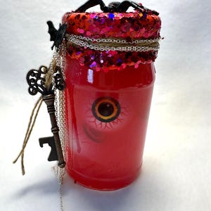 Può includere: Un barattolo di vetro rosso riempito con liquido rosso e un grafico di un grande bulbo oculare. Il coperchio è coperto di glitter rossi e sormontato da un ragno nero. Una catena d'argento, spago e una chiave scheletro in metallo pendono dal collo.