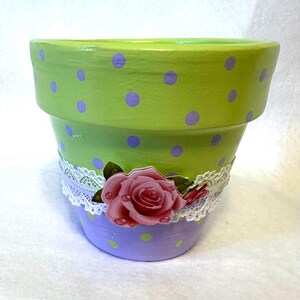 May include: A green and purple polka dot terracotta flower pot with a white lace trim and a pink rose embellishment.