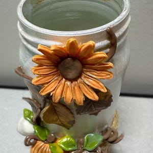 Op de afbeelding: Een witte keramische pot met een oranje zonnebloem en herfstdecoraties. De pot is versierd met een grote zonnebloem, pompoenen en bladeren. De pot heeft een rustieke, landelijke uitstraling.