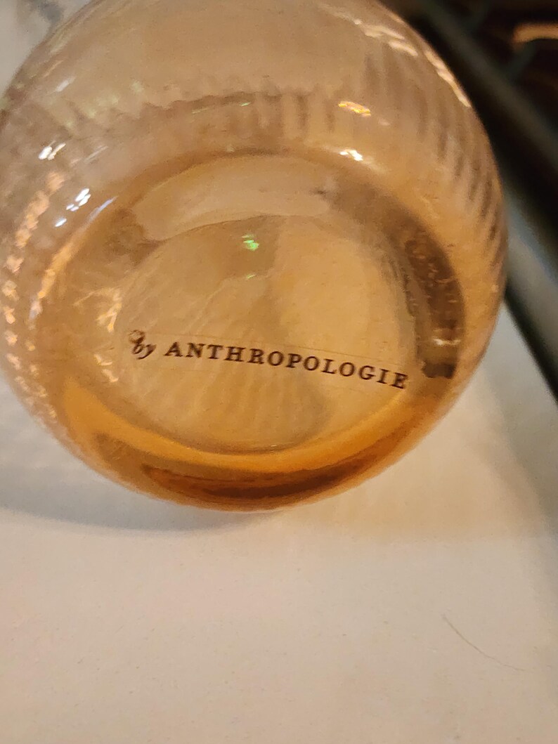 Puede incluir: Una botella de vidrio transparente con una superficie texturizada y el texto "by ANTHROPOLOGIE" impreso en la parte inferior.