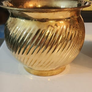 May include: A gold-colored, decorative planter with a spiral ribbed design. The planter has a rounded body, a flared rim, and a small, circular base. The metallic finish reflects light, creating a warm, inviting aesthetic. Ideal for small plants or decorative items.