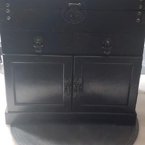 May include: A black wooden cabinet with two doors and a top drawer. The cabinet has ornate metal handles and hinges.