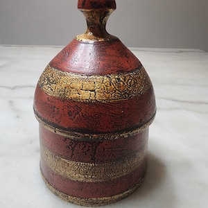 May include: A decorative, lidded jar with a red and gold color scheme. The jar has a rounded body with gold bands and a pointed top. The surface has a textured appearance, giving it a vintage or handcrafted feel.