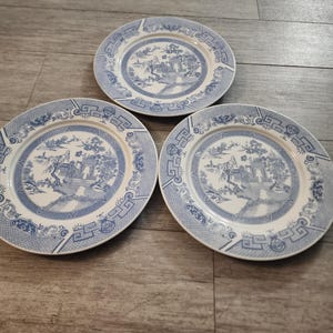 May include: Three blue and white ceramic plates with a traditional Chinese design. The plates have a white background with a blue border and a blue design in the center. The design features a pagoda, trees, and a bridge.