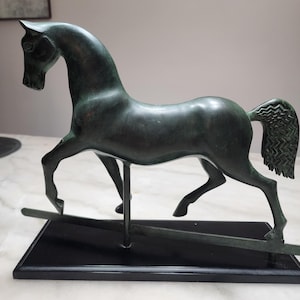 May include: A bronze-colored horse sculpture in a trotting pose. The horse has a sleek, curved body and a detailed tail. It is mounted on a black rectangular base with a metal support structure, suggesting a rocking horse design. The sculpture is a decorative item.
