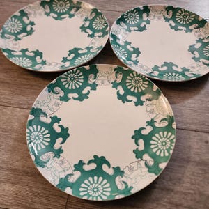 May include: Three white ceramic plates with a green and white floral design. The plates have a scalloped edge and are perfect for serving a meal.