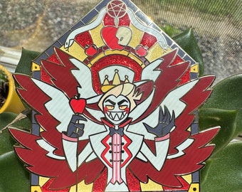 May include: A gold-coloured enamel pin featuring a cartoon character with red wings, a crown, and a red apple. The character is wearing a white suit with a pink tie and has a devilish grin. The pin is set against a red, gold, and white background.