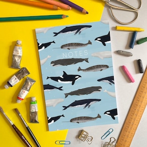 Può includere: Un quaderno blu con un'illustrazione di balene e narvali. Il quaderno ha la parola "NOTES" stampata in nero sulla copertina.