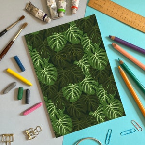 May include: A green notebook with a monstera leaf pattern and the word "NOTES" in gold lettering. The notebook is surrounded by art supplies, including paint tubes, pencils, and crayons.