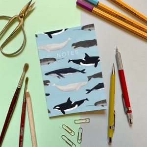 May include: A blue notebook with a whale pattern and the word "NOTES" printed on the front. The notebook is surrounded by pencils, pens, and paintbrushes on a light green surface.