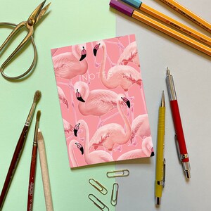 Op de afbeelding: Roze notitieboekje met het woord "NOTES" gedrukt op de omslag. Het notitieboekje is versierd met een patroon van roze flamingo's.