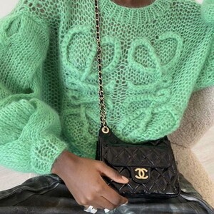 K&ouml;nnte beinhalten: Ein hellgr&uuml;ner, offen gestrickter Pullover mit Rundhalsausschnitt und strukturiertem Design auf der Vorderseite. Eine schwarze, gesteppte Umh&auml;ngetasche mit goldener Kette und goldenem Logo wird in der Hand gehalten.