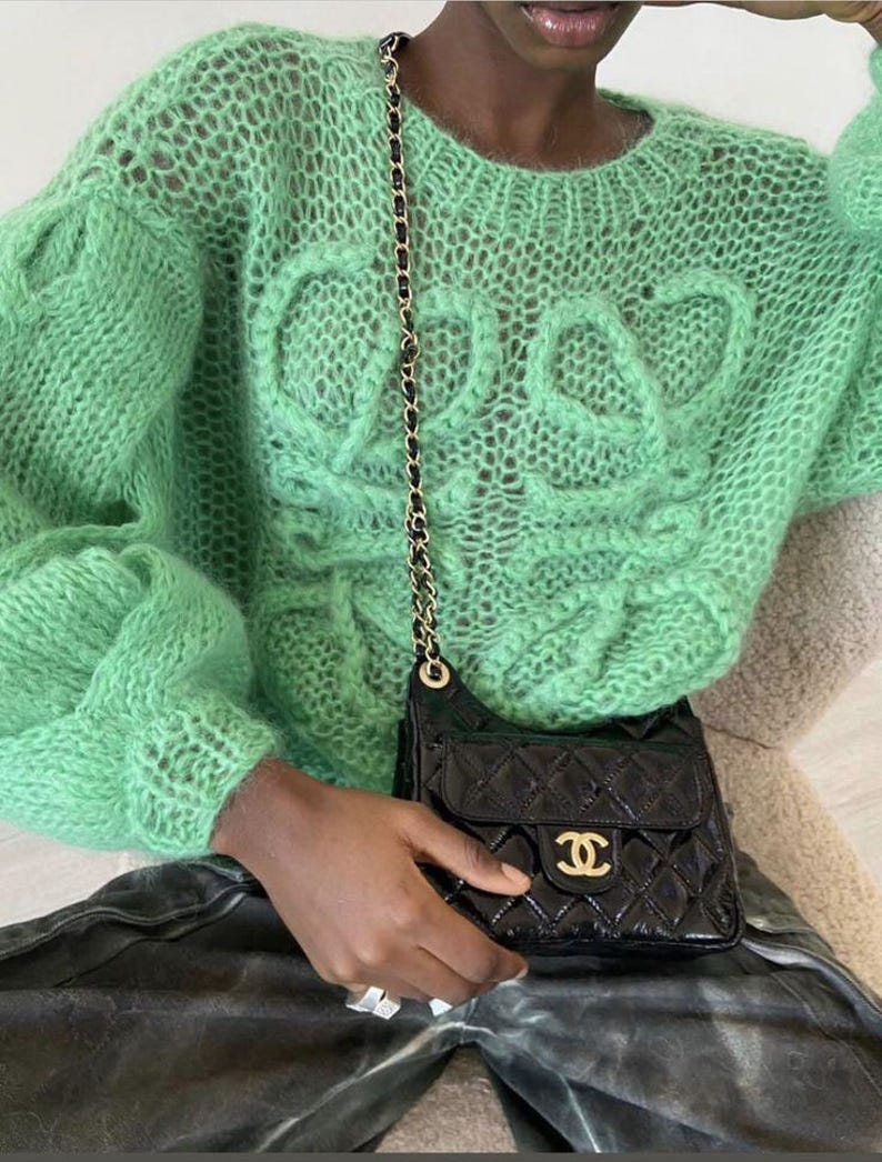 K&ouml;nnte beinhalten: Ein hellgr&uuml;ner, offen gestrickter Pullover mit Rundhalsausschnitt und strukturiertem Design auf der Vorderseite. Eine schwarze, gesteppte Umh&auml;ngetasche mit goldener Kette und goldenem Logo wird in der Hand gehalten.