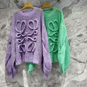 K&ouml;nnte beinhalten: Zwei Strickpullover h&auml;ngen an einer Metallstange. Einer ist helllila, der andere leuchtend gr&uuml;n. Beide Pullover haben ein gro&szlig;es, stilisiertes Design auf der Vorderseite. Die Pullover haben lange &Auml;rmel und einen runden Halsausschnitt.