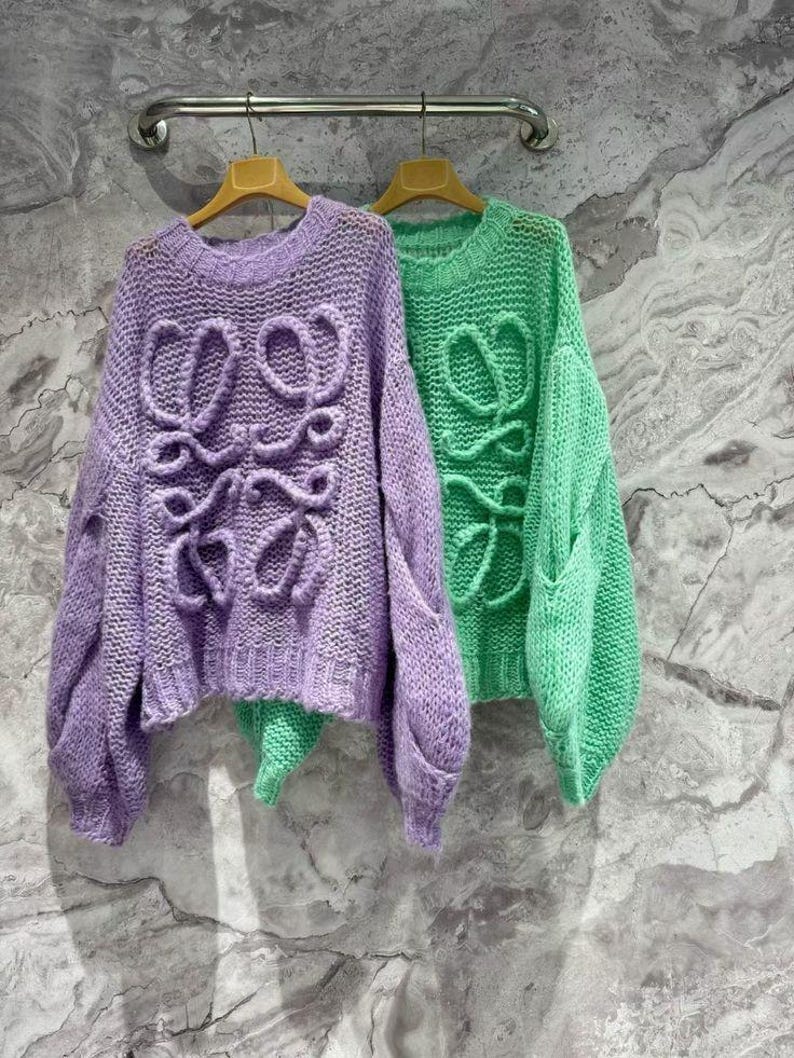 K&ouml;nnte beinhalten: Zwei Strickpullover h&auml;ngen an einer Metallstange. Einer ist helllila, der andere leuchtend gr&uuml;n. Beide Pullover haben ein gro&szlig;es, stilisiertes Design auf der Vorderseite. Die Pullover haben lange &Auml;rmel und einen runden Halsausschnitt.