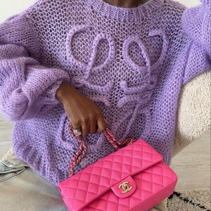 K&ouml;nnte beinhalten: Ein fliederfarbener, offen gewebter Pullover mit dem Wort "COS" auf der Vorderseite. Der Pullover hat lange, volumin&ouml;se &Auml;rmel. Eine leuchtend pinkfarbene, gesteppte Lederhandtasche mit goldener Kette und Verschluss wird in der Hand gehalten.