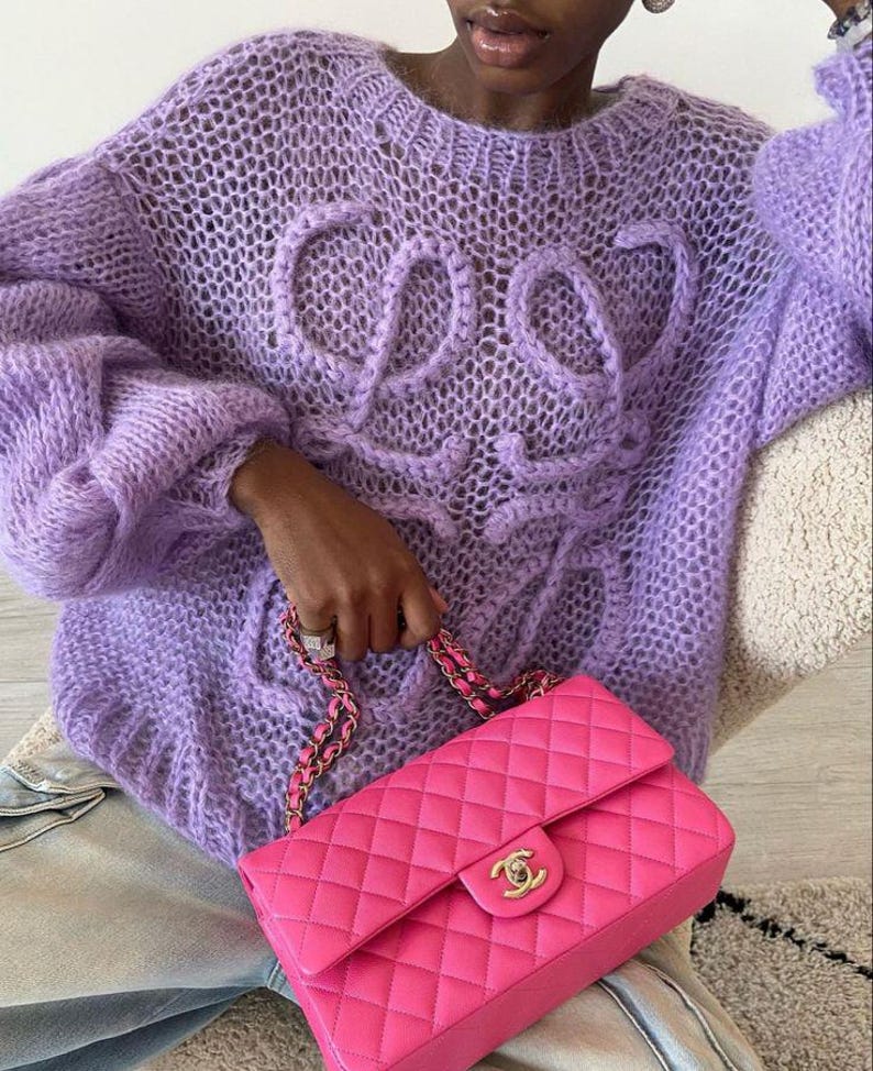 K&ouml;nnte beinhalten: Ein fliederfarbener, offen gewebter Pullover mit dem Wort "COS" auf der Vorderseite. Der Pullover hat lange, volumin&ouml;se &Auml;rmel. Eine leuchtend pinkfarbene, gesteppte Lederhandtasche mit goldener Kette und Verschluss wird in der Hand gehalten.