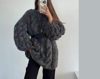 May include: A chunky, dark gray cable-knit sweater with oversized sleeves and a black belt cinching the waist. The sweater has a longline silhouette and is paired with black pants. The overall style is cozy and fashionable.