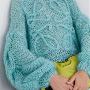 May include: A pale blue, open-knit jumper with a round neck and long, voluminous sleeves. The jumper has a textured design on the front. A yellow clutch is held in front of the jumper.