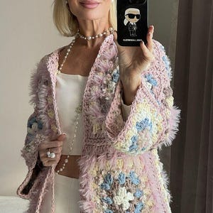 May include: A light pink crochet jacket with a square pattern and fluffy trim. The jacket features a mix of pastel colors, including blue, yellow, and white. The jacket is worn over a white top and pants.