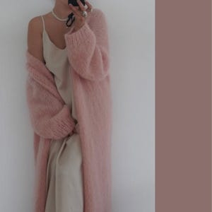 Pode incluir: Cardigã comprido e folgado rosa claro com uma textura macia e difusa. O cardigã é usado sobre um vestido de seda creme. As mangas são compridas e volumosas. O fundo é uma parede branca lisa com um sotaque castanho liso.
