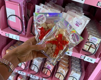May include: A pink vending machine filled with various packaged treats. A clear bag of white chocolate candies tied with a red ribbon is held in front. The machine displays prices and labels like "Chocolate Eggs" and "Mystery Bag."