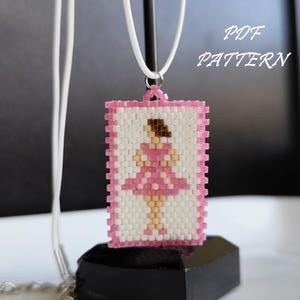Puede incluir: Un colgante de cuentas rosa y blanco con una bailarina con un vestido rosa. El colgante está colgado de un cordón blanco y está sobre un soporte negro. El texto "PDF PATTERN" es visible en la imagen.