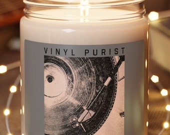 Puede incluir: Una vela blanca con una etiqueta gris que dice "Vinyl Purist" y una imagen en blanco y negro de un tocadiscos. La vela está sentada sobre un posavasos de madera y rodeada de luces blancas.