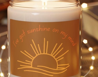 Puede incluir: Un tarro de vidrio para velas con una vela blanca en su interior. El tarro tiene una etiqueta marrón con un sol y el texto "I've got sunshine on my mind". La vela está sobre un posavasos de madera.